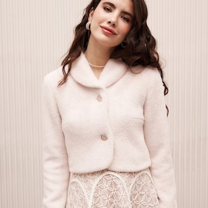 May include: A woman wearing a white fluffy cropped jacket with a collar and two buttons. She is also wearing a white lace skirt with a floral pattern.