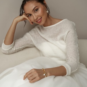 May include: A woman wearing a white sweater with pearl embellishments and a white skirt. She is wearing a gold chain bracelet with a pearl charm.