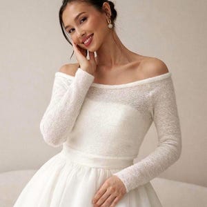May include: A white off-the-shoulder wedding dress with long sleeves. The dress features a fitted bodice and a full skirt. The sleeves and bodice have a textured, open-knit design. The bride is smiling and wearing gold earrings.