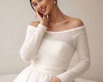 May include: A white off-the-shoulder wedding dress with long sleeves. The dress features a fitted bodice and a full skirt. The sleeves and bodice have a textured, open-knit design. The bride is smiling and wearing gold earrings.