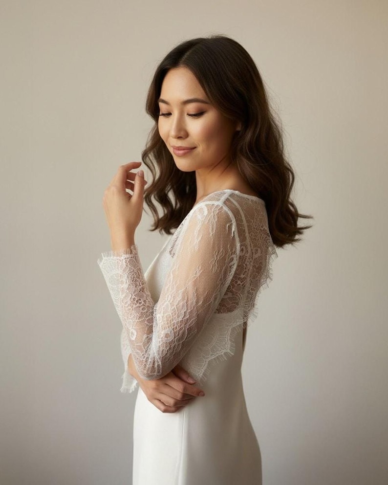 May include: A white wedding dress with long lace sleeves and a lace back. The dress has a simple, elegant design. The bride is looking down with a gentle expression.