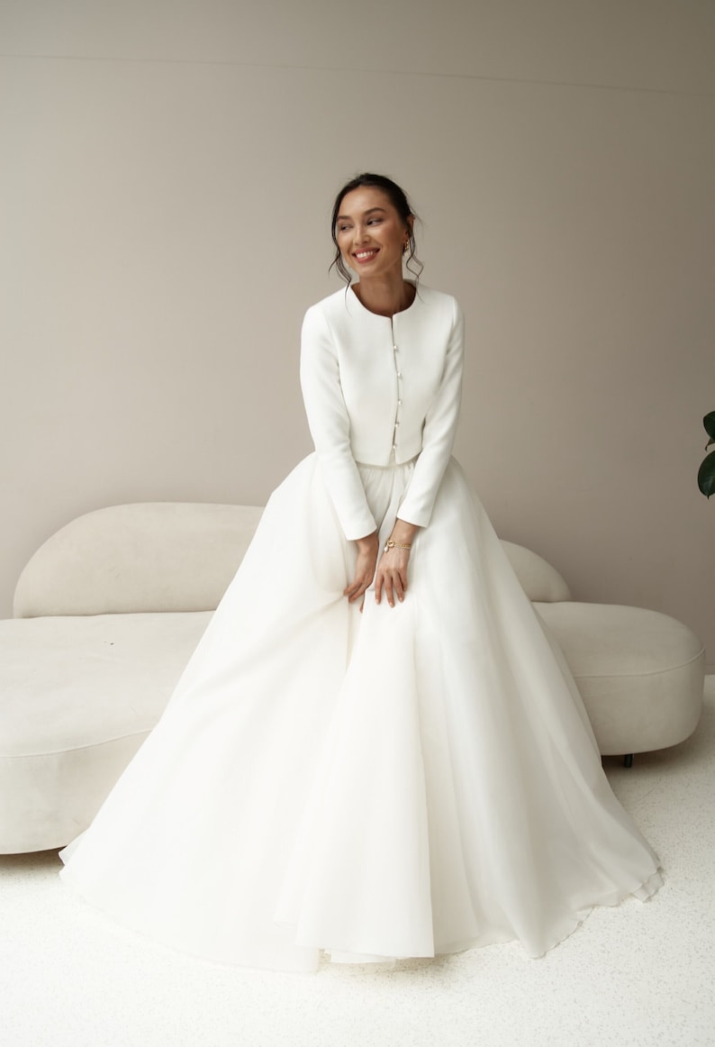 May include: A white bridal gown with a fitted bodice and a full skirt. The gown features a long-sleeved jacket with pearl buttons.
