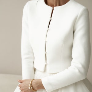May include: A white cropped jacket with long sleeves and pearl button closures. The jacket is made of a soft, smooth fabric.