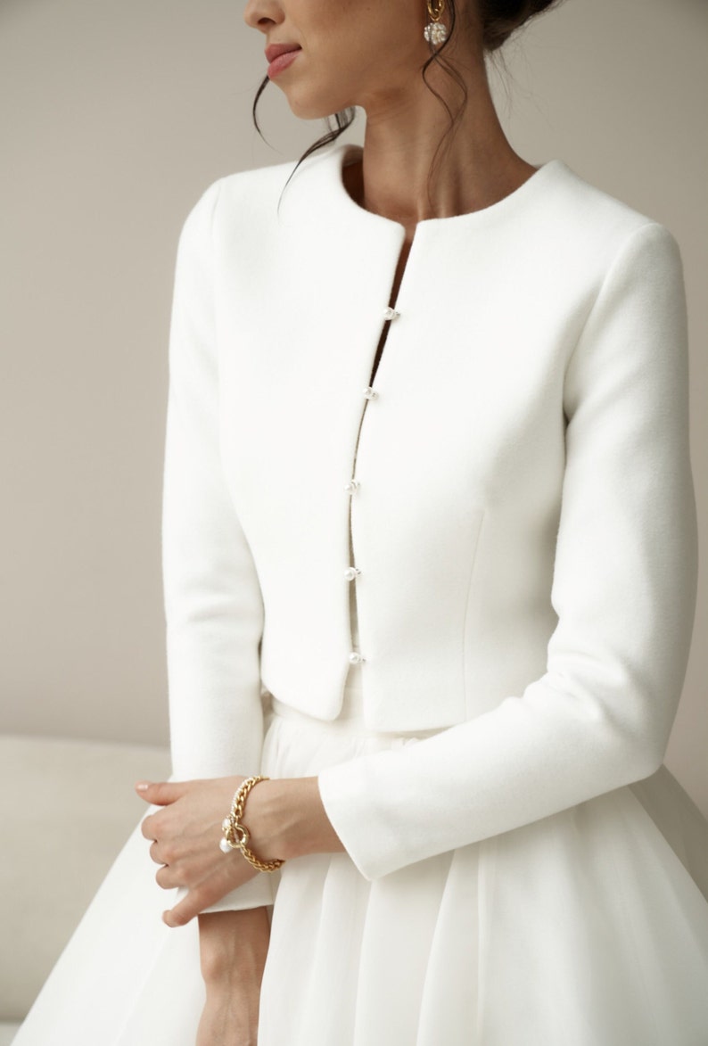 May include: A white cropped jacket with long sleeves and pearl button closures. The jacket is made of a soft, smooth fabric.