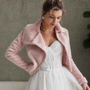 Könnte beinhalten: Hellrosa Kunstfelljacke mit breitem Kragen und langen Ärmeln. Die Jacke wird über einem weißen Brautkleid mit herzförmigem Ausschnitt und Tüllrock getragen. Das Kleid hat ein eng anliegendes Oberteil und einen fließenden Rock.