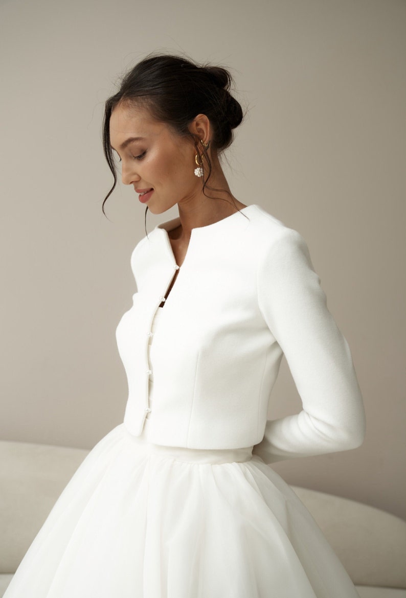 May include: A white bridal jacket with long sleeves and a button-up front. The jacket is cropped and has a simple, elegant design.