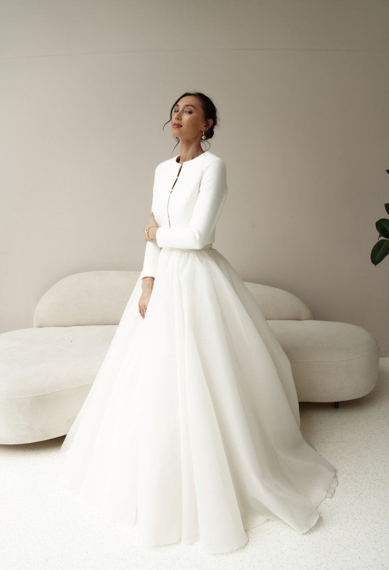 May include: A white bridal gown with a full skirt and a matching white jacket. The jacket has long sleeves and a button closure.