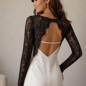 May include: A white dress with a black lace back and long sleeves. The dress features a low back with a thin white strap and a V-neckline. The sleeves are made of intricate black lace.