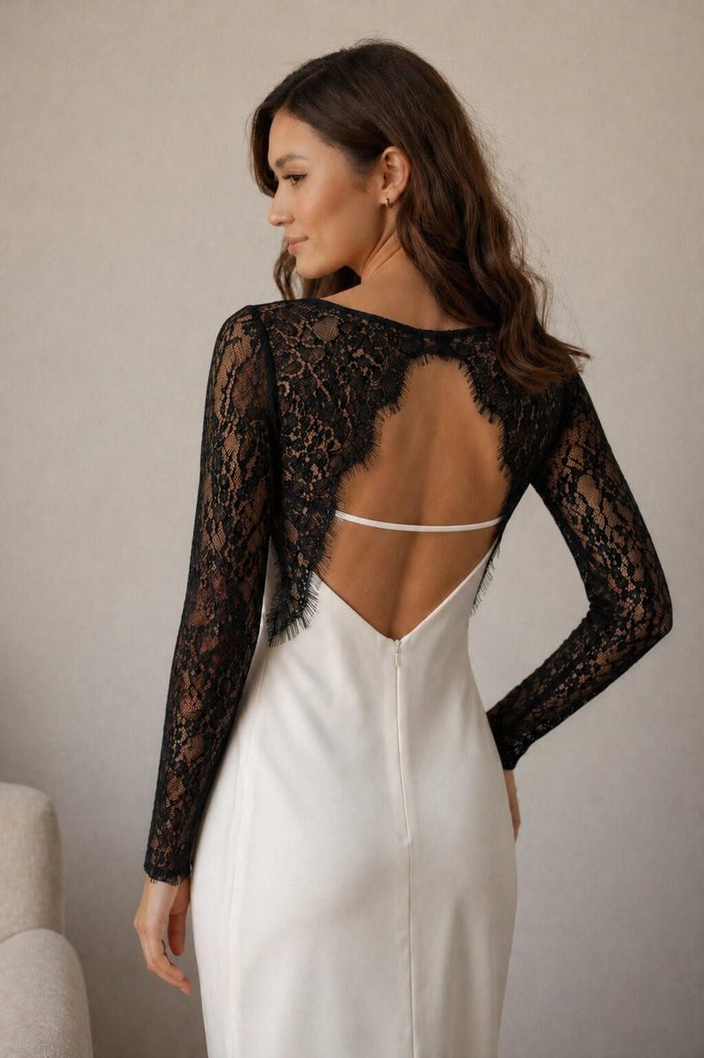 May include: A white dress with a black lace back and long sleeves. The dress features a low back with a thin white strap and a V-neckline. The sleeves are made of intricate black lace.