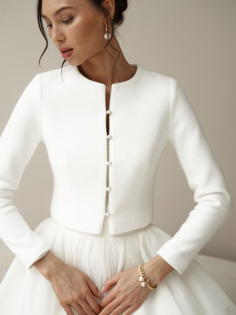 May include: A white bridal jacket with long sleeves and a round neckline. The jacket has a single row of pearl buttons down the front.