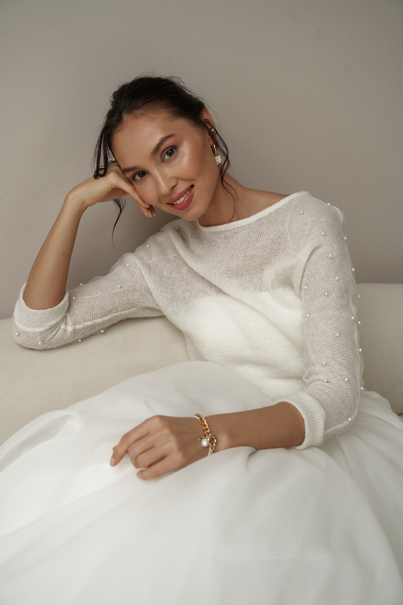 May include: A woman wearing a white sweater with pearl embellishments and a white skirt. She is wearing a gold chain bracelet with a pearl charm.