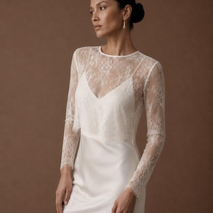 May include: A floor-length white gown featuring a sleeveless satin slip dress base with a sheer, long-sleeved lace overlay. The overlay has a round neckline and intricate floral lace patterns that extend down the sleeves to the wrists.