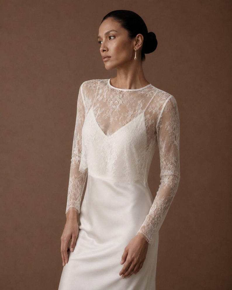 May include: A floor-length white gown featuring a sleeveless satin slip dress base with a sheer, long-sleeved lace overlay. The overlay has a round neckline and intricate floral lace patterns that extend down the sleeves to the wrists.