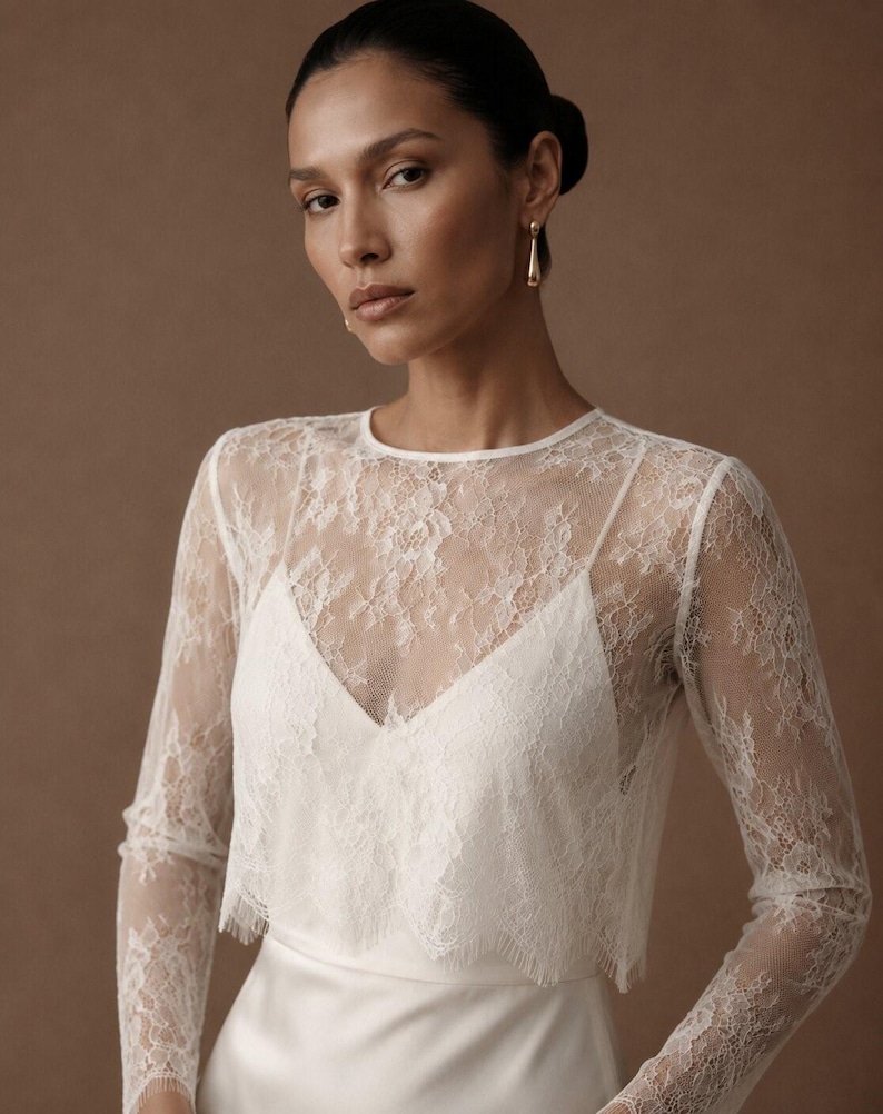 May include: A white lace long-sleeve top featuring a delicate floral pattern and a scalloped hemline. The garment has a round neckline and is layered over a white camisole with a V-neckline, creating a textured and semi-sheer appearance.