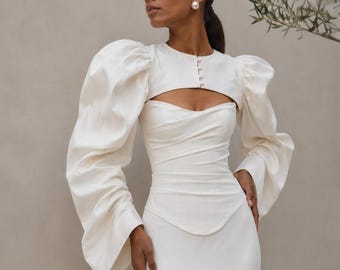 May include: An ivory-colored dress featuring a bodice with a sweetheart neckline and a cropped overlay top with long, voluminous puff sleeves. The overlay includes a round neckline with a vertical row of small buttons and a cutout detail.