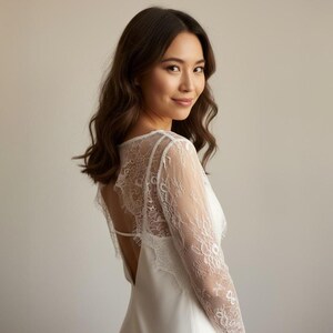 May include: A white wedding dress with a lace back and sleeves. The dress has a low back and a simple, elegant design. The model is looking over her shoulder with a slight smile.