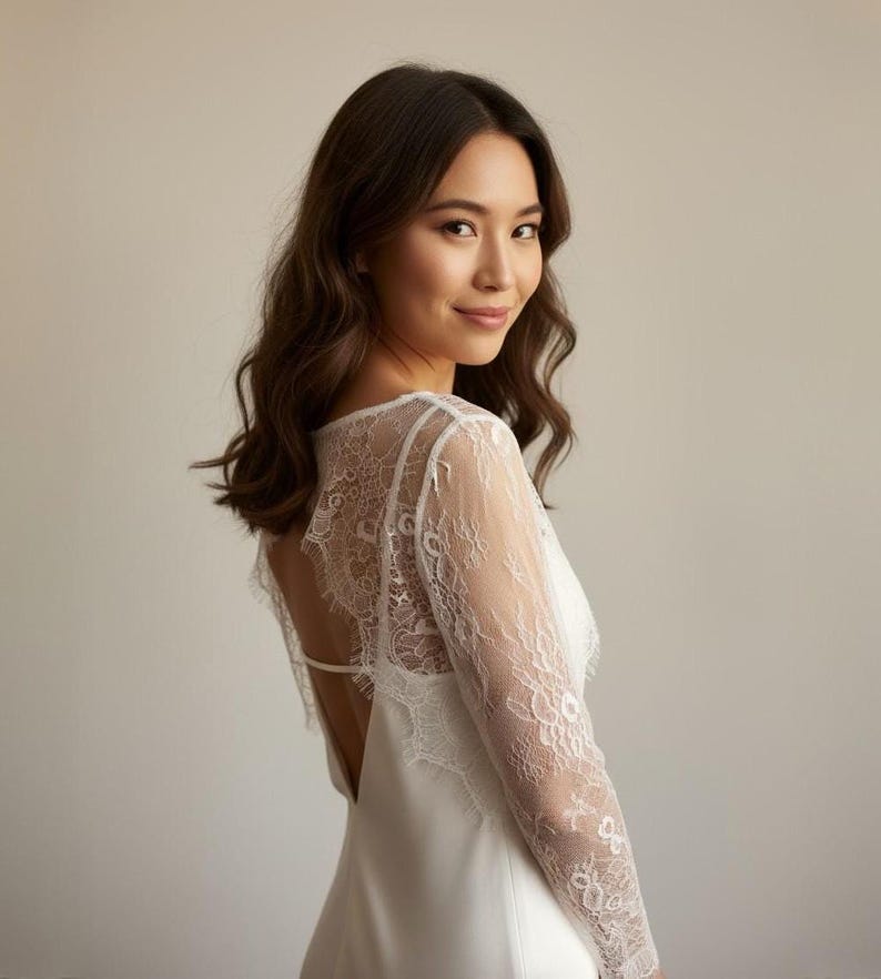 May include: A white wedding dress with a lace back and sleeves. The dress has a low back and a simple, elegant design. The model is looking over her shoulder with a slight smile.