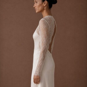 May include: A floor-length white gown featuring long, sheer lace sleeves and a lace bodice overlay. The garment has a high neckline and a deep, open back design that exposes the upper back area, transitioning into a smooth, solid white skirt.