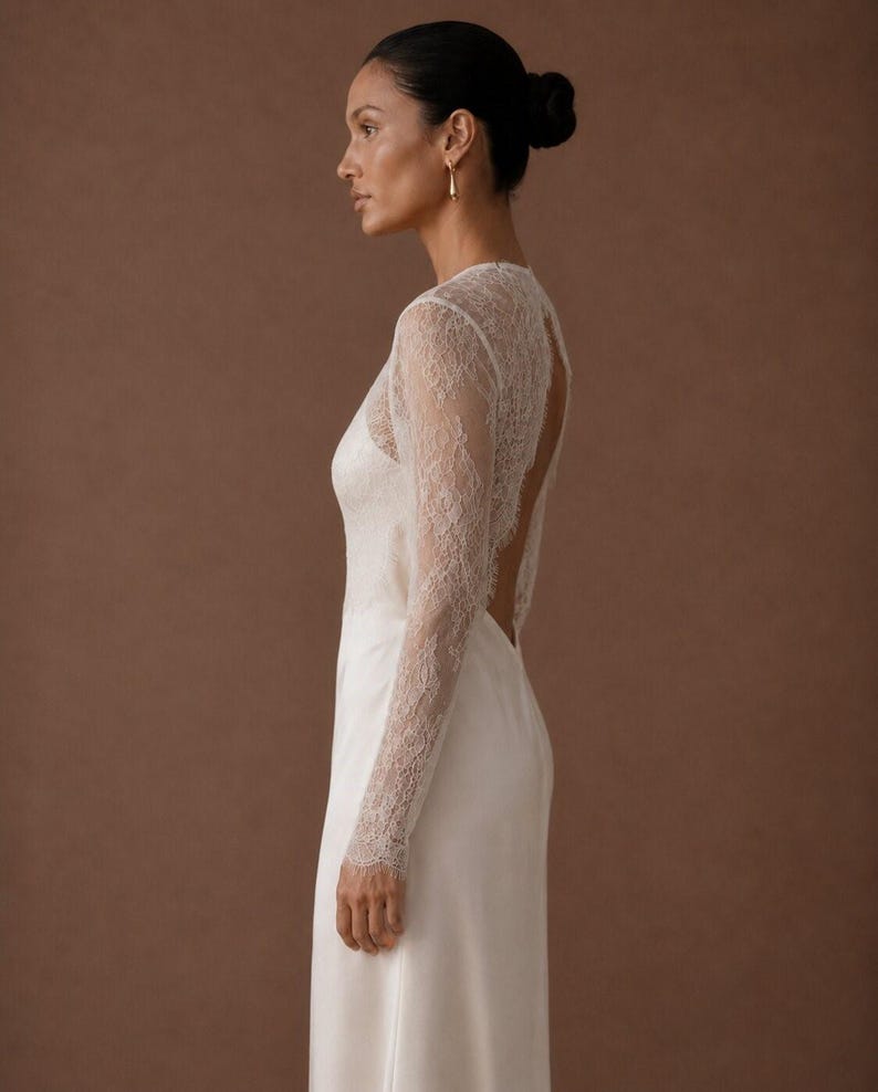 May include: A floor-length white gown featuring long, sheer lace sleeves and a lace bodice overlay. The garment has a high neckline and a deep, open back design that exposes the upper back area, transitioning into a smooth, solid white skirt.