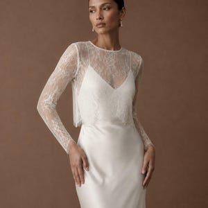 May include: A floor-length white satin dress featuring a sheer lace overlay with long sleeves and a round neckline. The bodice has a V-neckline underneath the lace, and the skirt is made of a smooth, solid white satin fabric that falls straight.
