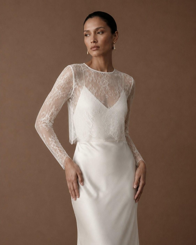 May include: A floor-length white satin dress featuring a sheer lace overlay with long sleeves and a round neckline. The bodice has a V-neckline underneath the lace, and the skirt is made of a smooth, solid white satin fabric that falls straight.