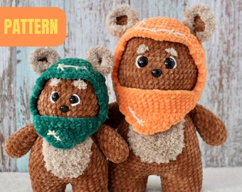 May include: Two crocheted plush toys in the shape of a brown Ewok with a furry face and large ears. The larger Ewok is wearing an orange hooded scarf and the smaller Ewok is wearing a green hooded scarf. The toys are standing on a white surface.