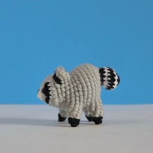 May include: A small, handcrafted crochet raccoon toy made with light grey yarn. It features black yarn details around the eyes, black-tipped paws, and a distinctive striped tail with alternating black and white yarn rings. The figure stands on four legs.
