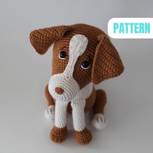 May include: A crocheted dog toy in shades of brown and white. The dog features large, drooping ears, a white face, and black button eyes. The word "PATTERN" is displayed in a blue box. The dog is in a sitting position.