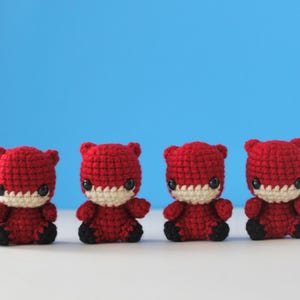 May include: Four red crocheted figures with white faces and black eyes. The figures are sitting in a row on a white surface.