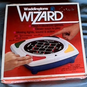 May include: A box for the Waddingtons Wizard electronic game. The box is red with white text and a picture of the game board. The game board is white with blue and red accents. The game board has a grid with lights and buttons. The box says "Plays 4 electronic games, Eleven ways to play, Moving lights, sound & action".