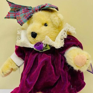 May include: A plush teddy bear wearing a purple velvet dress with a white lace collar, a green bow, and a purple flower. The bear has a plaid bow on its head and black shoes.