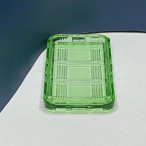 May include: A rectangular, light green glass dish with a grid pattern. The dish has a slightly raised edge and is likely used for serving or display. The glass has a smooth, reflective surface.