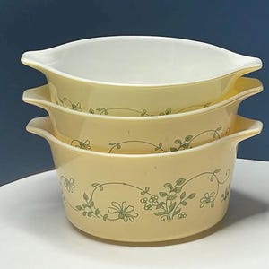 May include: A stack of three pale yellow glass bowls with integrated side handles. Each bowl features a white interior and a delicate green botanical pattern of vines and small flowers encircling the exterior. The bowls are nested together.
