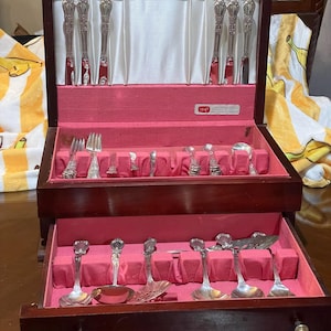 May include: A wooden silverware chest with a red velvet interior, holding various silver utensils. The chest is open, revealing knives, forks, spoons, and serving pieces. The text "1847" is visible on the inside.