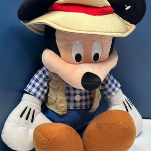 May include: A plush Mickey Mouse toy wearing a safari hat with a red band, a tan vest, and a blue and white checkered shirt. The toy has large black ears, white gloves with black lines, and orange feet.