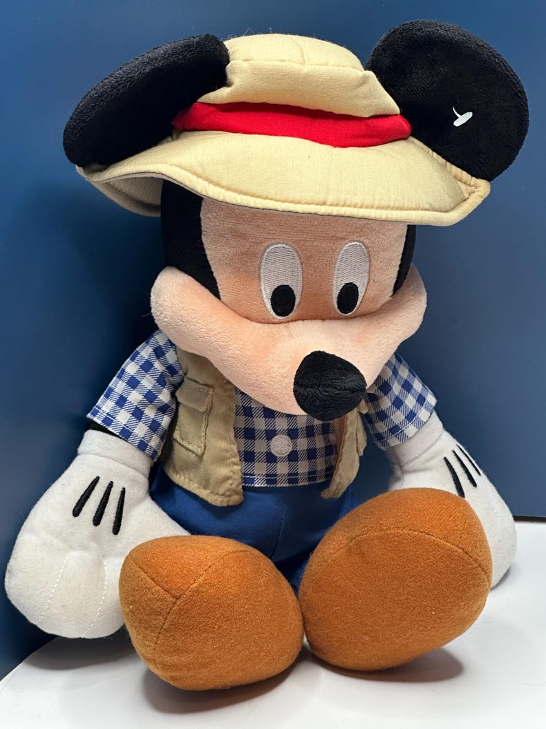 May include: A plush Mickey Mouse toy wearing a safari hat with a red band, a tan vest, and a blue and white checkered shirt. The toy has large black ears, white gloves with black lines, and orange feet.