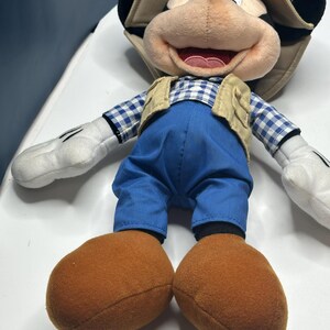 May include: A plush Mickey Mouse doll wearing a tan safari hat, a tan vest, blue pants, and a blue and white checkered shirt. The doll has large brown feet and white gloves. The doll is lying on a white surface.