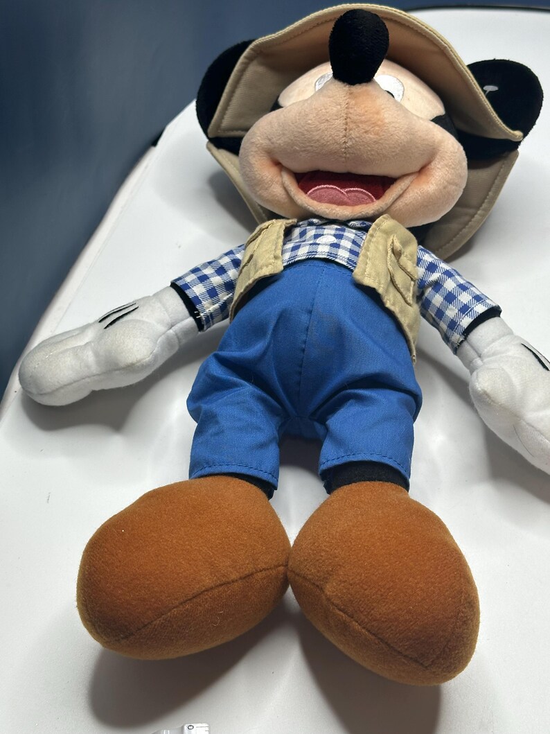 May include: A plush Mickey Mouse doll wearing a tan safari hat, a tan vest, blue pants, and a blue and white checkered shirt. The doll has large brown feet and white gloves. The doll is lying on a white surface.