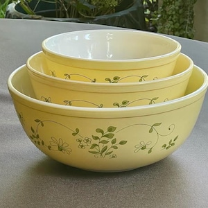 May include: A set of three nested mixing bowls in a pale yellow color. Each bowl features a repeating green botanical pattern of delicate vines, leaves, and small flowers encircling the exterior. The interior of the bowls is a smooth, solid white.