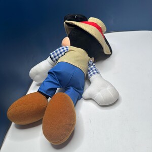 May include: A plush Mickey Mouse toy wearing a tan vest, blue pants, and a straw hat with a red band. The toy has large brown shoes, white gloves, and a black head with large ears. The toy is lying on its side.