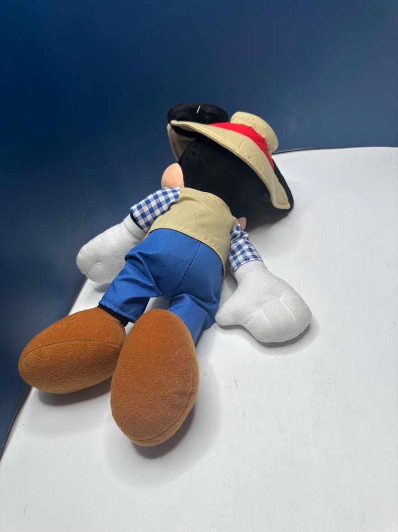 May include: A plush Mickey Mouse toy wearing a tan vest, blue pants, and a straw hat with a red band. The toy has large brown shoes, white gloves, and a black head with large ears. The toy is lying on its side.