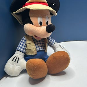 May include: A plush Mickey Mouse toy wearing a safari hat with a red band, a tan vest, and a blue and white checkered shirt. The toy has large black ears, white gloves, and orange feet.