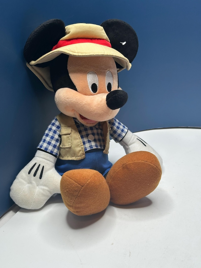May include: A plush Mickey Mouse toy wearing a safari hat with a red band, a tan vest, and a blue and white checkered shirt. The toy has large black ears, white gloves, and orange feet.