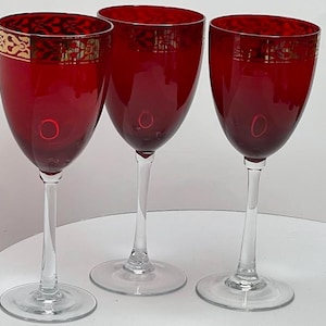 May include: Three red wine glasses with gold trim around the top. The glasses are clear and have a stemmed base.