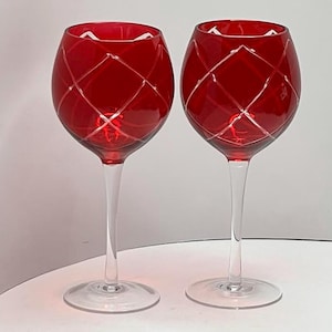 May include: Two red wine glasses with clear stems and bases. The red bowls have a diamond pattern etched into the glass. The glasses are on a white surface.
