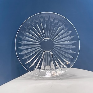 May include: A clear glass serving plate featuring a sunburst pattern of radiating ridges that extend from a smooth circular center to the outer rim. The plate is displayed upright on a thin metal stand, showcasing its transparent, reflective surface.