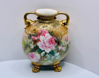 May include: A vintage porcelain vase with a round body, three gold feet, and two handles. The vase is decorated with a floral pattern of pink roses and green leaves, accented with gold details.