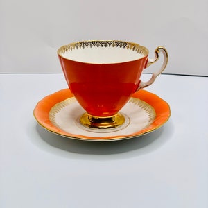 May include: A vintage teacup and saucer set in a bright orange color with gold trim. The cup has a white interior and a delicate gold design around the rim.