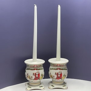 May include: A pair of white ceramic candle holders with a Christmas theme. Each holder features a Santa Claus design with a red suit and white beard. The holders have a gold trim and are decorated with green holly leaves and red berries.
