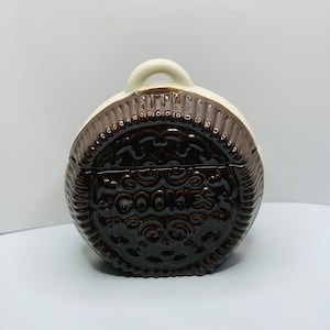May include: A brown and white ceramic cookie jar shaped like a giant chocolate chip cookie. The jar has a handle on top and says "COOKIES" in the center.